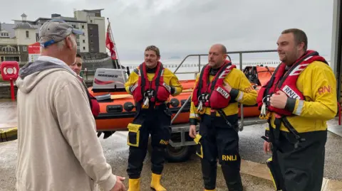 Mr Grant is standing to the left facing three RNLI crew members who are wearing waterproofs and life jackets. There is a life boat behind them and the beach at Weston-super-Mare.