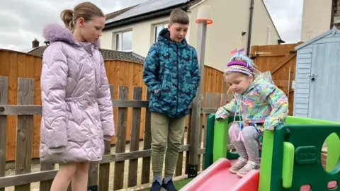 Ella is sat on a slide in her garden while her brother and sister look on. 