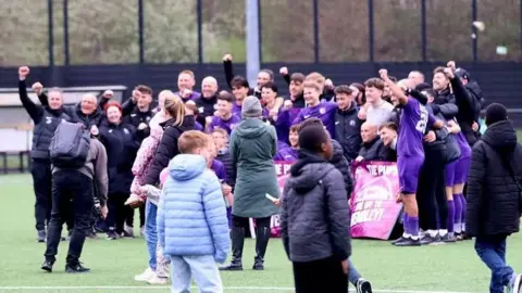 Those White Lines A crowd of people are gathered together on a football pitch pumping their fists in the air in celebration. They are holding a large banner that cannot be made out because people are in front of it.  