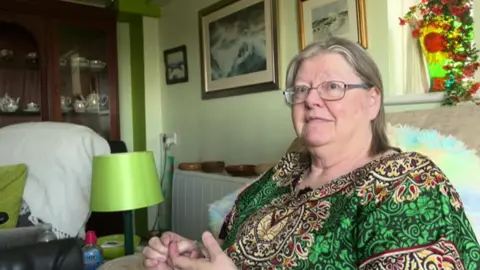 Maggie sits in an armchair looking at the camera, but sits side on. She is wearing a colourful patterned shirt with lots of green and has glasses.