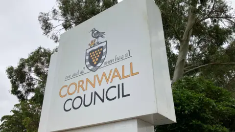 BBC White sign reads "One and all Cornwall Council". Green trees are behind the sign. 