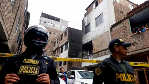 Two police officers guard a cordoned off area, which has several apartment blocks that are about four storeys high. One of them is blackened by fire.