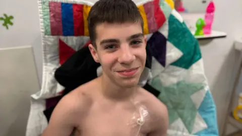 Finley sitting upright in a hospital bed. He has a medical tube attached to his chest. He is smiling at the camera. On the bed behind him is a multi-coloured patchwork blanket. Finley has dark brown straight hair.