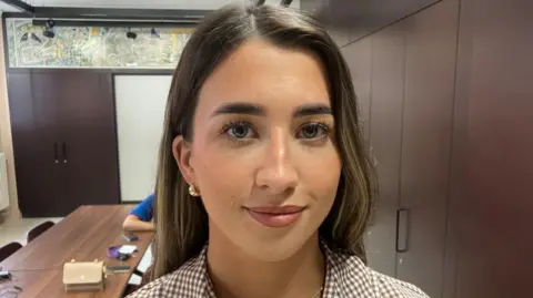 A young woman, Rhian McCrory. She is wearing a plaid top and had brunette hair. Behind her are cupboards and a table.