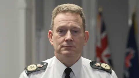 Ben-Julian Harrington is wearing a white shirt and black tie with black and gold epaulettes on his shoulders. He has short brown hair and looks at the camera with a serious expression. Behind him are a Union Jack flag and a navy Essex Police flag.