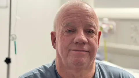 Nigel Hamnett is sitting in the hospital. He has closely cut grey hair and is wearing a light blue top. 