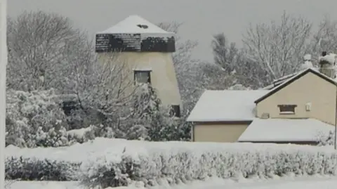 Keith Jones Snow covers a windmill style home plus neighbouring houses. It is also resting on the branches of the trees and bushes.