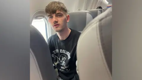 Tyler Kerry, who has short brown hair and stubble, sitting in an aeroplane seat. He is wearing a black t-shirt. The picture has been taken by someone in the row of seats in front of him.