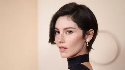 Gracie Abrams poses on the red carpet of the 2026 Oscars, in a black dress with a choker, and sparkling diamond earrings.