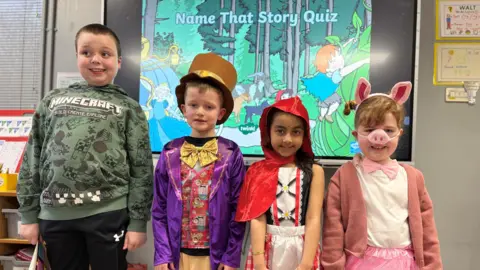 Four children in a classroom dressed in costume for World Book Day.   Jack, who is tall and has short dark hair, is wearing a green jumper with a Minecraft logo.  Mason is wearing a Willie Wonka outfit which consists of a bright purple jacket, a brown top hat and a gold bow tie.  Izabella, who has long, dark hair, is wearing a red cape as Red Riding Hood.  Scarlett has blonde hair and is wearing a pink snout, pink ears on a headband and a pink cardigan and skirt. 