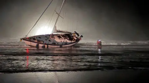 Polzeath Coastguard An overturned white sailing yacht on a flat sandy beach beach at night, illuminated from behind. The mast - without sails - is pointing about 35 degrees, towards land. The tide is half out, and a person in red stands halfway between the shore and boat ankle-deep in water.