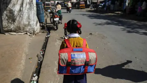 A girl wearing a school uniform - light brown sweater over a white shirt and navy-blue shirt - carries a schoolbag coloured red, blue and grey on her back as she walks on a road in India's western state of Maharashtra on a sunny day. 