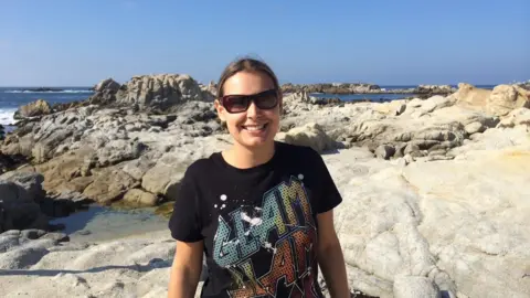 Tarryn Baird standing in front of rocks on a bench in front of the sea wearing a black T-shirt that says "glam slam".