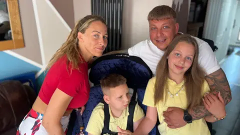 A family of four hugs each other and looks towards the camera. On the left is a woman with blonde hair in a red top, next to her in a wheelchair in a yellow polo shirt is a boy, and on the right is a man in a white t-shirt hugging a girl in a yellow polo shirt