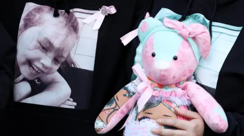 Reuters A woman holds a pink and light blue teddy bear to her chest, covering a T-shirt with a black and white picture of Olivia Pratt-Korbel smiling on it. Another woman wear the same T-shirt alongside her. Only their torsos are in shot