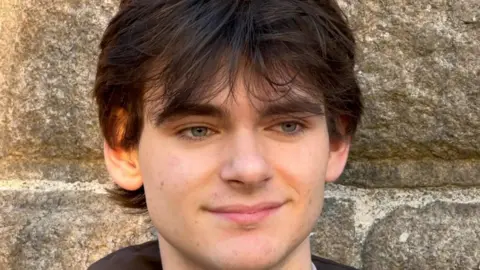 A young man with dark brown, slightly tousled hair, standing against a textured stone wall. 