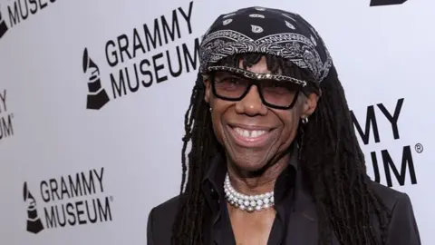 Nile Rodgers, wearing a black bandana on top of his head, a pearl necklace, and dark glasses, stands smiling in front of a white wall of the Grammy Museum. He is wearing a black jacket.