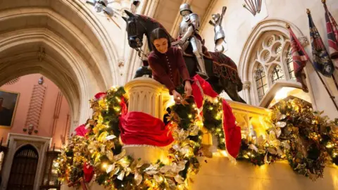 Royal Collection Enterprises Limited 2025 A woman leaning over the Grand Staircase is making adjustments to the garlands draped around it.