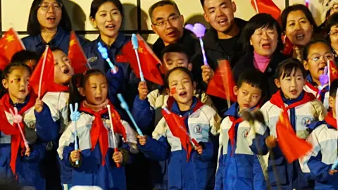 BBC/Xiqing Wang Crianças em uniformes escolares agitam bandeiras e cantam enquanto se despedem da tripulação