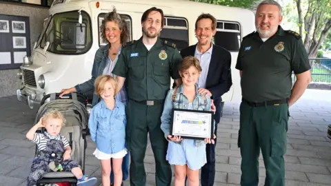 London Ambulance Service From left: Gabriel in a pushchair, Anouk, mother Nadja, call handler Matthew Hawkins, Farah, father Scott, and deputy director of 999 operations James Eden