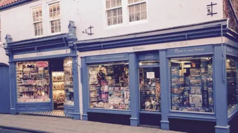 A quaint, traditional shopfront with two large display windows and a central entrance, painted in a muted blue colour. The windows are filled with colourful items including books, magazines, toys, and puzzles. Above the windows, signage reads ‘Holmans’. The building has white upper walls with sash windows and decorative black iron brackets.