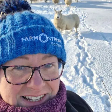 Sally Crowe Sally takes a selfie in the snow. She is wearing brown tortoiseshell glasses, a purple woolly jumper, a dark jacket, and a bright blue bobble hat which says "Farm Strong Scotland". There is a sheep behind her.