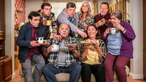 BBC Eight people gathered around a sofa in a living room. Some of them are carrying a selection of mugs, sandwiches and cakes. Two people are on the sofa, two on the arms of the sofa and four are standing behind with two of them reaching out to the plates of food