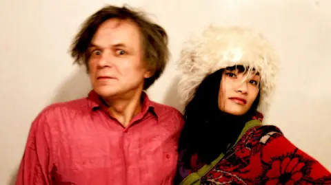 Artist Sergei Ivanov wearing a bright textured shirt stands beside his partner Tszman Chan dressed in patterned clothing. She is wearing a large, fluffy light‑coloured hat and has a scarf draped across the shoulder. Both individuals pose against a plain, softly lit background.