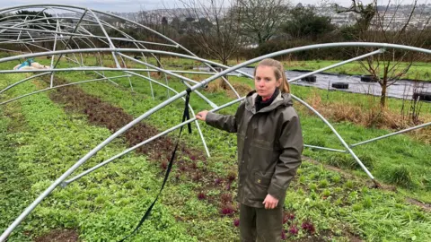 Abi Gwynn stands next to the bent out of shape frame of tunnel normally used to grow fruit and vegetables. She is wearing a waterproof coat.