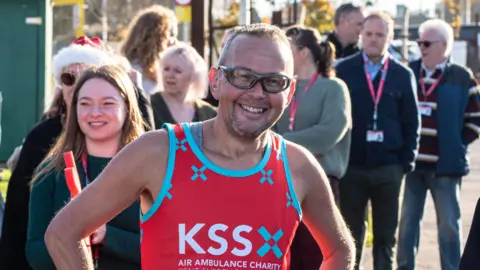 A man wearing sunglasses and a red KSS running vest. Behind him is a crowd of people and he is smiling at the camera. 