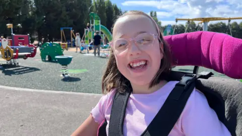 Girl with glasses smiles at the camera - she is strapped into a motorised wheelchair and behind her is a playpark with children playing