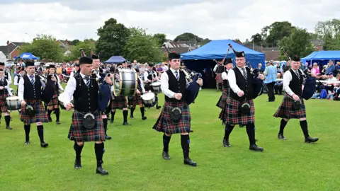 Pacemaker Press A group are holding pipes and drums and walking across grass. Spectators are watching. The sky is cloudy. Marquees are up. 