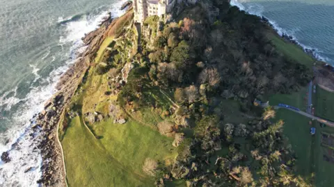 PA Media The picture shows an aerial view of a rocky island surrounded by the sea, with waves crashing against the shoreline. At the top of the island sits a large historic castle-like building made of stone, perched on elevated ground. 
The island itself is covered in a mix of manicured green lawns and dense patches of trees. Some areas of the trees appear damaged or fallen from strong winds or a recent storm.