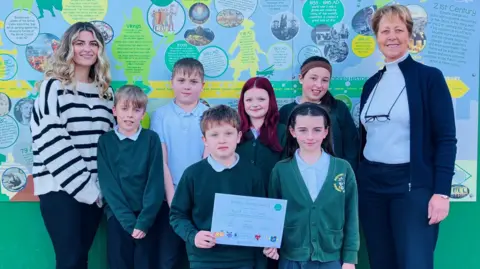 East Riding of Yorkshire Council Six children hold a certificate to celebrate their school being given a health award. Mel Wilson, the school safeguarding lead, stands on the left of the image - she is wearing a blue and white striped jumper and has wavy blonde hair. Councillor Victoria Aitken is standing on the right of the image. 