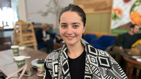 A woman with her dark hair tied back, wearing a black and white diamond patterned jacket over a black top, sitting at a cafe table.