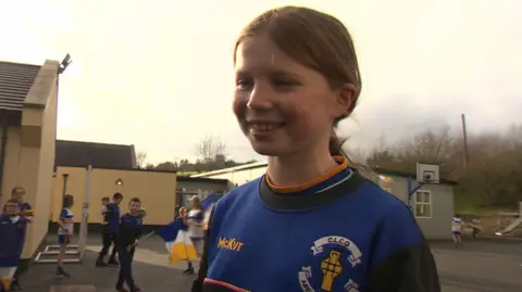 A young girl with fair long hair tied back in a ponytail.
She is in a school playground and is wearing a blue GAA sports top.