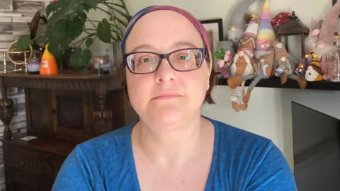 A woman in a blue t-shirt with glasses and a pink headband. A plant in the background on a shelf with lots of toys on too. There are three picture frames on the wall behind. 