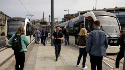 People at a tram stop in Edinburgh with two trams waiting on either side of the platform
