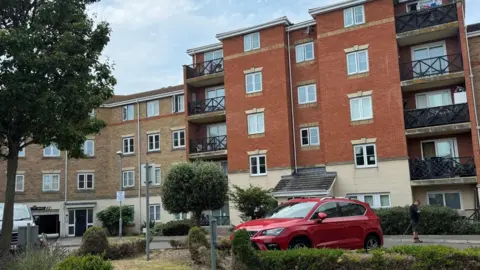 Elliot Deady/BBC A block of flats, with a red car parked outside beside a small garden