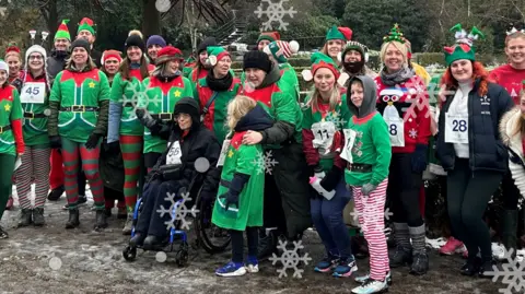 A group photo of a previous Mental Elf event, with participants dressed up as elves standing on a snowy and icy field. One woman is sat in a wheelchair on the front row and the runners have numbers on their chest.