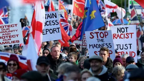A large crowd gathers at a protest in Crowborough carrying Union flags and banners. Visible signs include messages reading "Stop the Madness" and "Not Racist". People fill the street in the foreground and background.