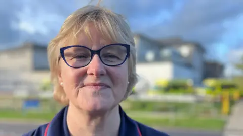 Head and shoulders of a lady with large navy rimmed glasses and short blonde hair in a bob looking directly at the camera, wearing a navy nurses uniform with red piping - a hospital with waiting ambulances is in the background