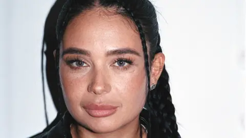 A girl with blue grey eyes, black braided hair and a black leather jacket in front of a white background.