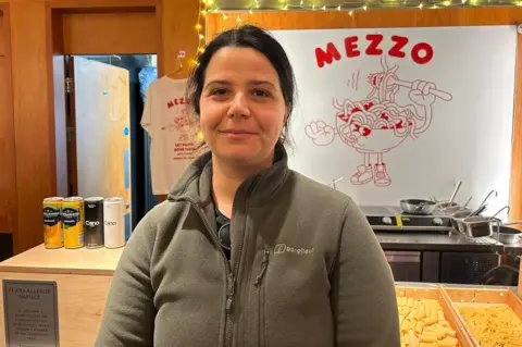 Alice Di Chiara, wearing a grey fleece sees at the camera. Behind her is pasta, saucepans, Italian drinks and a Mezzo sign with red letters and a bowl of pasta with legs holding a fork