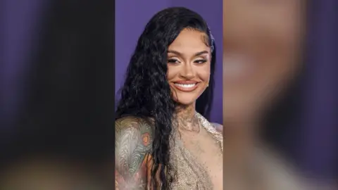 Kehlani, with long black hair and shoulder tattoos, smiles to camera, against a purple background
