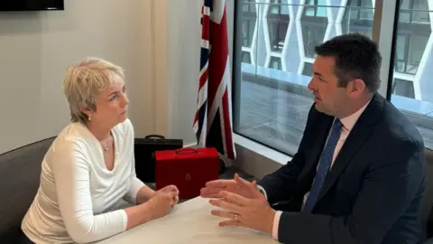Shaun Davies MP Shaun Davies MP and health minister Karin Smyth talking at a table in Parliament with a union flag on a flag pole behind them, plus brief cases