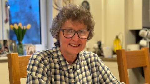 A woman in a yellow blue check shirt sat in a kitchen. She has grey hair and black glasses