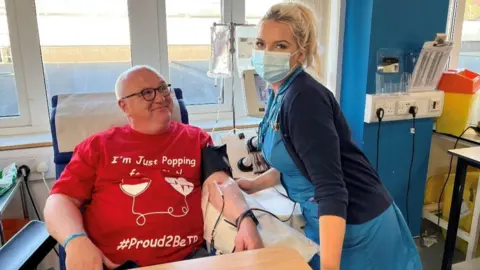 Somer Elliott A blood donor lying down with a tube in his arm. He is wearing a red t-shirt saying he is 'proud to be a donor'. Somer is standing next to him wearing a mask and checking he is well. 