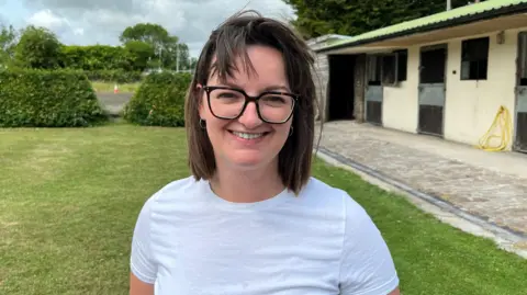 A young woman with medium-length brown hair and wearing glasses, smiles at the camera whilst standing on grass with a hedge and dilapidated single-story building in background.