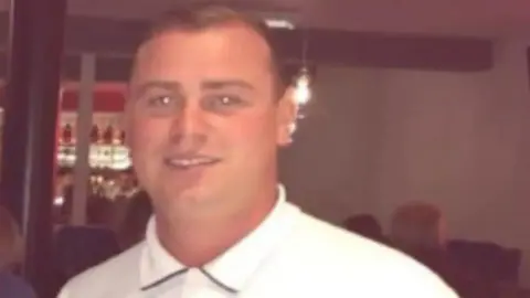 Rico Burton smiles wearing a white polo shirt while stood in a bar. The backs of other people's heads can be seen behind him. 